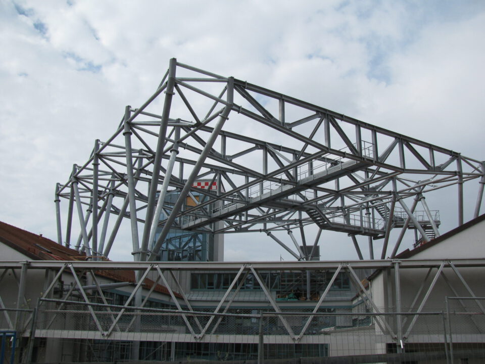 Großes Stahltragwerk mit zahlreichen miteinander verbundenen Trägern und Stützen, das eine komplexe geometrische Struktur bildet; im Inneren ist eine Treppe erkennbar, im Hintergrund sind weitere Gebäude und ein bewölkter Himmel zu sehen.