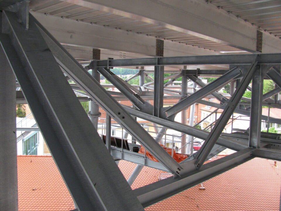 Nahaufnahme eines Stahltragwerks mit mehreren Trägern und Fachwerkkonstruktionen in Dreiecksform, vermutlich Teil eines Gebäudes im Bau; im Hintergrund sind ein Ziegeldach und umliegende Gebäude durch die Struktur hindurch sichtbar.