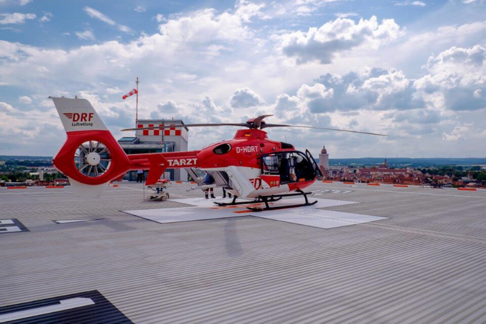 Ein rot-weißer Notarzt Hubschrauber mit der Kennung „D-HDRT“ und der Aufschrift „DRF Luftrettung“ steht auf einem Hubschrauberlandeplatz; im Hintergrund ist eine Stadtlandschaft unter einem teils bewölkten Himmel zu sehen.