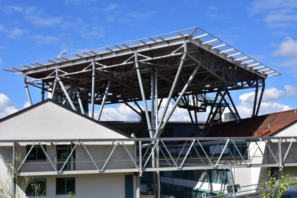 Ein großes, erhöhtes Metalltragwerk ruht auf mehreren Stahlstützen über dem Hauptgebäude mit weißen Wänden und Fenstern; ein brückenartiger Steg mit Metallgeländer verbindet verschiedene Gebäudeteile, im Hintergrund ist ein teils bewölkter Himmel zu sehen.