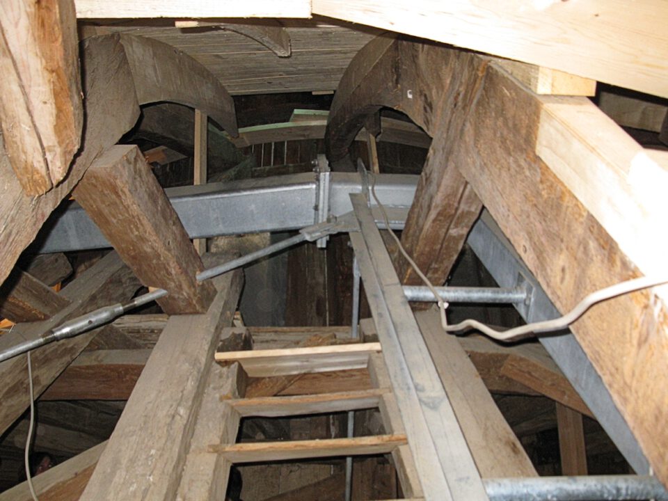 Innenansicht eines hölzernen Gebäudes mit großen Balken, Metallstützen und Rohren; in der Bildmitte steht eine senkrechte Leiter, die zu höheren Ebenen führt, die Konstruktion wirkt rustikal und unfertig.