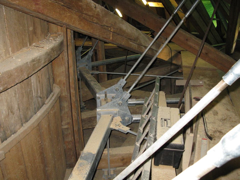 Nahaufnahme einer hölzernen Innenkonstruktion mit Balken, Metallverstrebungen und einem großen zylindrischen Holzelement links; Werkzeuge und Materialien wie ein Werkzeugkasten liegen verstreut, die Umgebung wirkt staubig und im Bau oder in Wartung.