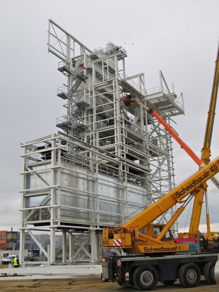 Das Bild zeigt ein großes Industriegebäude im Bau mit mehreren Ebenen aus Metallträgern und Treppen, wobei ein Kran einen Arbeiter in einem orangefarbenen Korb zu einer oberen Ebene hebt; am Boden sind weitere Arbeiter zu sehen, der Himmel ist bewölkt.