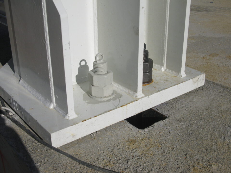 Das Bild zeigt eine Nahaufnahme einer Stahlstütze mit Grundplatte, die mit großen Schrauben – eine weiß, eine schwarz – auf einem Betonfundament verankert ist.