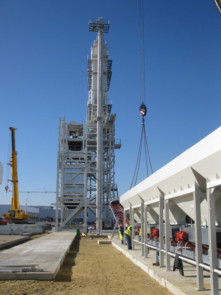 Das Bild zeigt eine Baustelle mit einer großen Metallstruktur. In der Mitte, einem gelben Kran links, mehreren Arbeitern mit Sicherheitsausrüstung rechts sowie einem herabhängenden Haken an Seilen vor blauem Himmel.