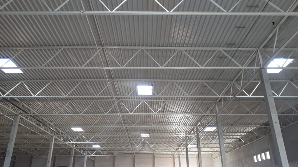 Das Innere einer großen Industriehalle mit Decke aus Wellblech, getragen von weiß gestrichenen Stahlträgern; mehrere rechteckige Oberlichter lassen Tageslicht einfallen, und vertikale Stützen gliedern den Raum.