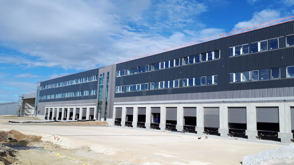 Großes Industriegebäude mit moderner Fassade, zwei Etagen und mehreren Laderampen, umgeben von einer Baustelle mit Erdhaufen und Baumaterialien unter leicht bewölktem Himmel.