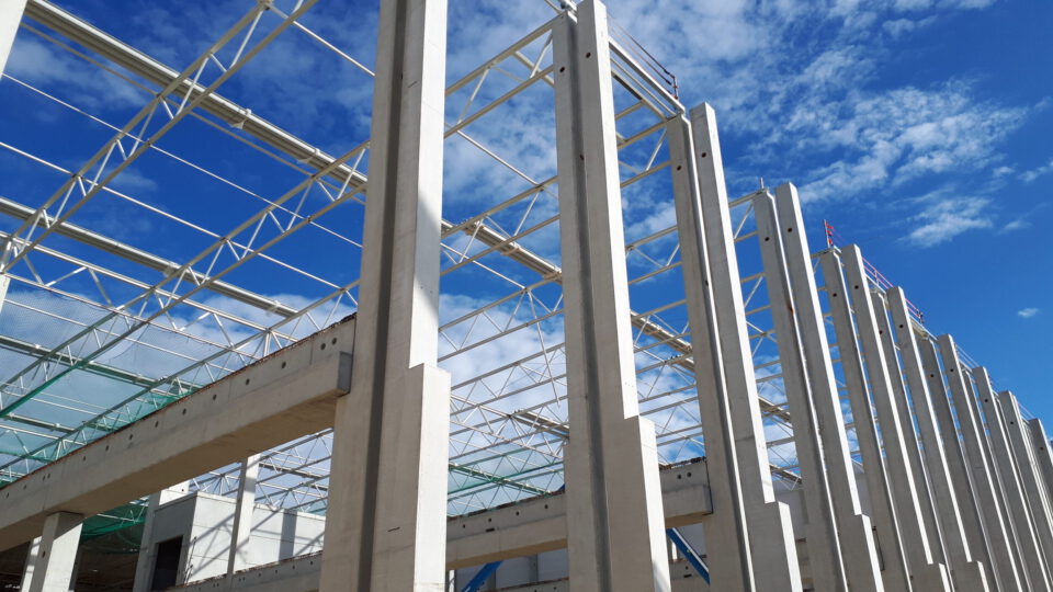 Baustelle eines großen Gebäudes mit hohen Betonsäulen und Stahlträgern, die das Grundgerüst bilden, unter leicht bewölktem Himmel.