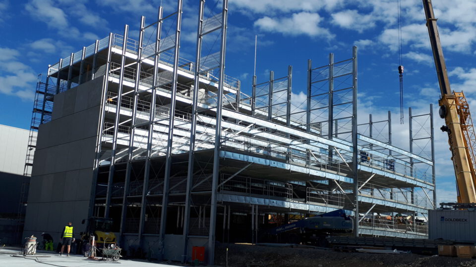 Baustelle mit einem mehrstöckigen Stahlgerüstbau, mehreren senkrechten Trägern und Plattformen, einem Kran am rechten Bildrand sowie Arbeitern und Baumaschinen im unteren Bereich unter teils bewölktem Himmel.