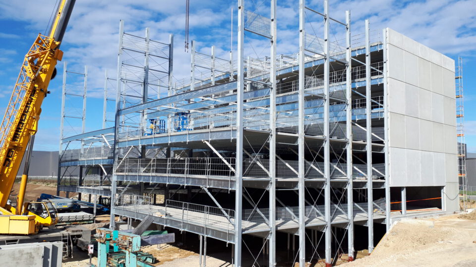 Baustelle mit einem mehrstöckigen Stahlbau im Rohbau, einem gelben Kran auf der linken Seite sowie verschiedenen Baumaschinen und Materialien rund um das Gelände unter leicht bewölktem Himmel.