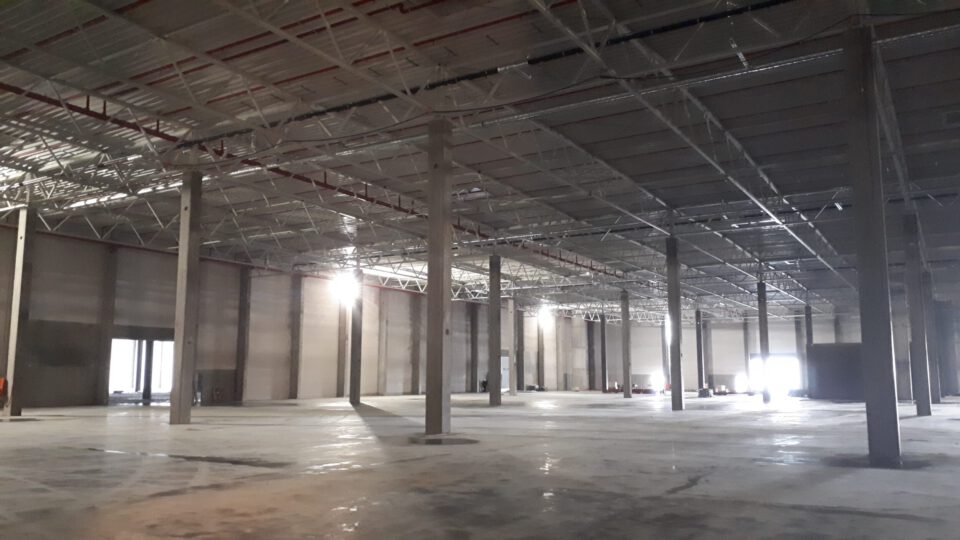 Innenraum einer großen, leeren Industriehalle mit hohen Decken, zahlreichen Betonsäulen, poliertem Betonboden sowie Lichtquellen durch Fenster und künstliche Beleuchtung.