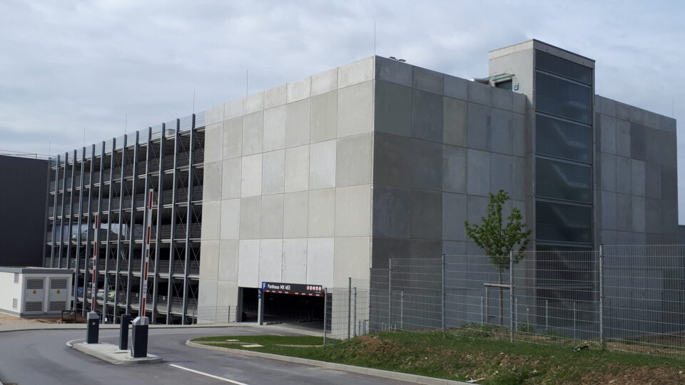 Mehrstöckiges Parkhaus mit mehreren Ebenen, einem verglasten Treppen- oder Aufzugsschacht auf der rechten Seite, Schrankenanlage am Eingang und einem Schild über der Einfahrt.