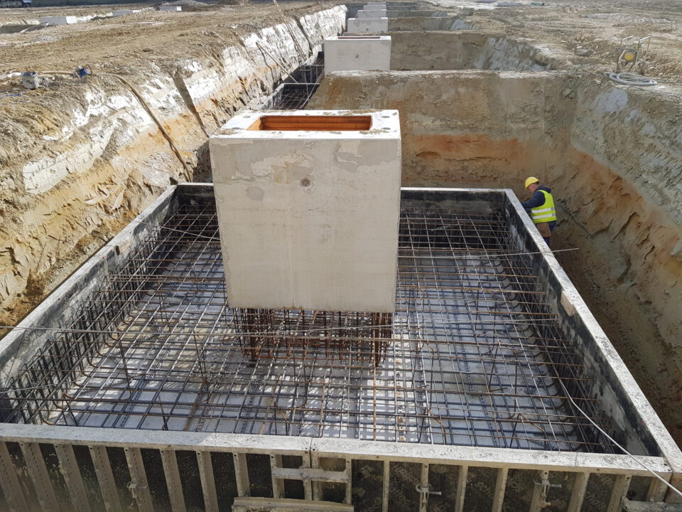 Eine Baustelle mit vorbereiteten Betonfundamenten, mehreren rechteckigen Fundamentkörpern mit Bewehrungsgittern und einem Bauarbeiter mit gelbem Helm und Warnweste, der in einer der Fundamentgruben arbeitet; umgeben von Erdreich und Aushubwänden.