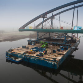 Großer Lastkahn auf einem Fluss, beladen mit Baumaschinen und Materialien; unter einer Brücke positioniert, die sich im Bau oder in Wartung befindet, mit mehreren Arbeitern und schwerem Gerät an Bord, umgeben von nebliger Landschaft.