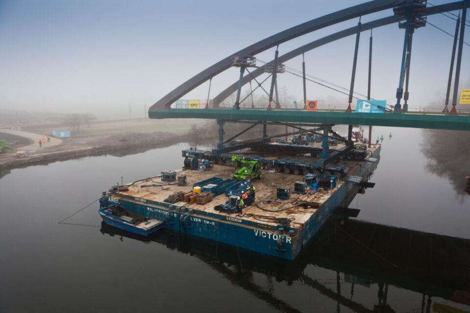 Großer Lastkahn auf einem Fluss, beladen mit Baumaschinen und Materialien; unter einer Brücke positioniert, die sich im Bau oder in Wartung befindet, mit mehreren Arbeitern und schwerem Gerät an Bord, umgeben von nebliger Landschaft.