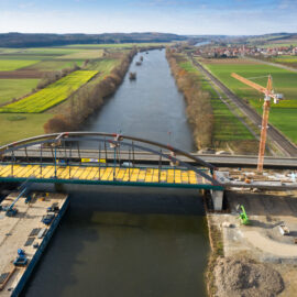 Im Bau befindliche Bogenbrücke über einen Fluss mit gelben Arbeitsplattformen auf der Oberseite; rechts steht ein Kran, im Hintergrund sind Felder, Straßen und eine kleine Ortschaft zu sehen.