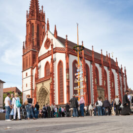 Das Bild zeigt eine große, reich verzierte Kirche mit rot-weißer Fassade und hohem Turm, vor der sich eine Menschenmenge versammelt hat; neben dem Eingang steht ein geschmückter hoher Pfahl unter leicht bewölktem Himmel.