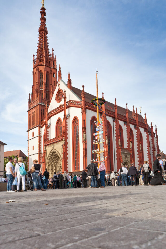 Das Bild zeigt eine große, reich verzierte Kirche mit rot-weißer Fassade und hohem Turm, vor der sich eine Menschenmenge versammelt hat; neben dem Eingang steht ein geschmückter hoher Pfahl unter leicht bewölktem Himmel.