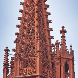 Das Bild zeigt die filigrane Spitze eines gotischen Bauwerks aus rötlich-braunem Stein mit zahlreichen Verzierungen wie Spitzbögen, Fialen und Maßwerk über mehrere Ebenen hinweg.