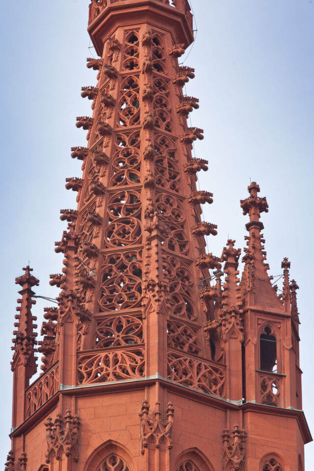 Das Bild zeigt die filigrane Spitze eines gotischen Bauwerks aus rötlich-braunem Stein mit zahlreichen Verzierungen wie Spitzbögen, Fialen und Maßwerk über mehrere Ebenen hinweg.