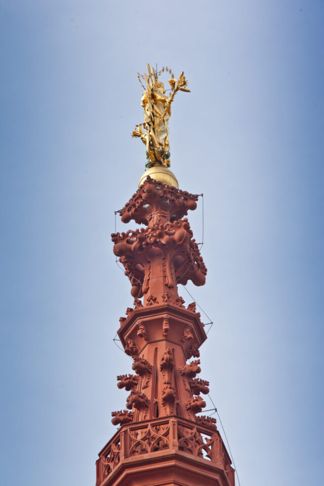 Das Bild zeigt die Spitze eines hohen, kunstvoll gestalteten Bauwerks aus rötlich-braunem Material mit filigranen Verzierungen und einer goldenen Statue einer Figur mit einem Gegenstand in der Hand vor klarem, blauem Himmel.