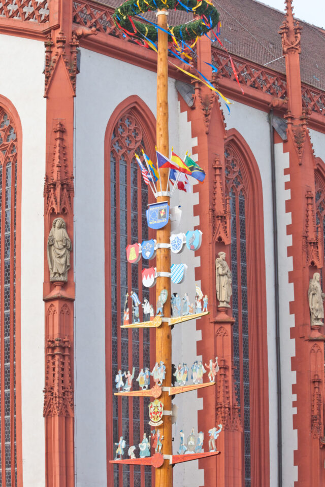Das Bild zeigt einen hohen, bunt geschmückten Holzpfahl mit Wappen, Fahnen und Figuren vor einem historischen Gebäude mit rot-weißer Fassade und verzierten architektonischen Details.