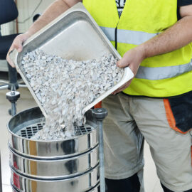 Das Bild zeigt eine Person in gelber Sicherheitsweste und beigefarbener Hose, die Kies aus einem Metallbehälter in ein Siebstapelgerät schüttet, zur Analyse der Korngrößenverteilung.