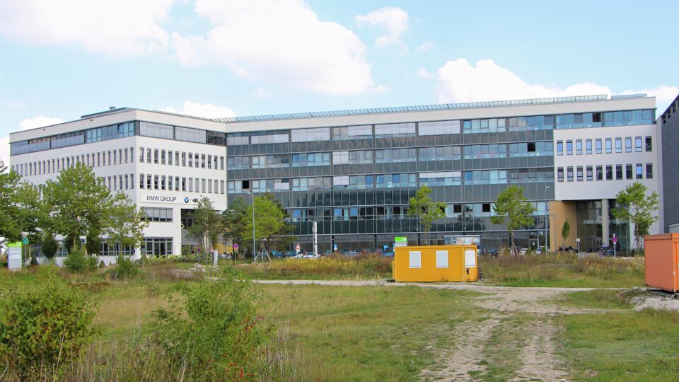 Ein modernes Bürogebäude mit mehreren Etagen und großen Fenstern; davor befinden sich Bäume, Grünflächen sowie ein gelber und ein orangefarbener Container, unter einem teilweise bewölkten Himmel.
