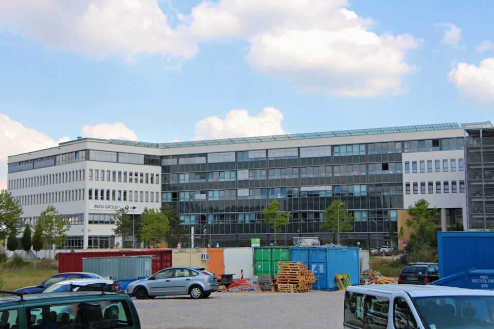 Ein großes, modernes Bürogebäude mit Glasfassade und der Aufschrift „BMW GROUP“ steht im Hintergrund; davor befinden sich geparkte Autos, bunte Container, Holzpaletten, Bäume und verstreute Baumaterialien unter einem teilweise bewölkten Himmel