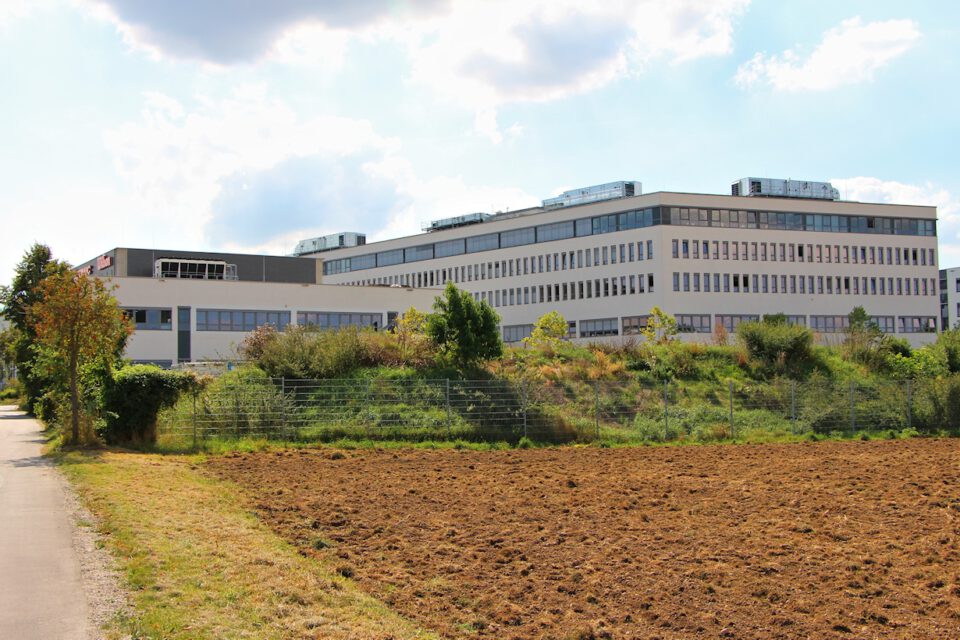 Ein großes, modernes Gebäude mit mehreren Etagen steht hinter einem Feld mit umgepflügtem Boden; links verläuft ein von Bäumen und Büschen gesäumter Weg, darüber ein teilweise bewölkter Himmel mit blauen Abschnitten.