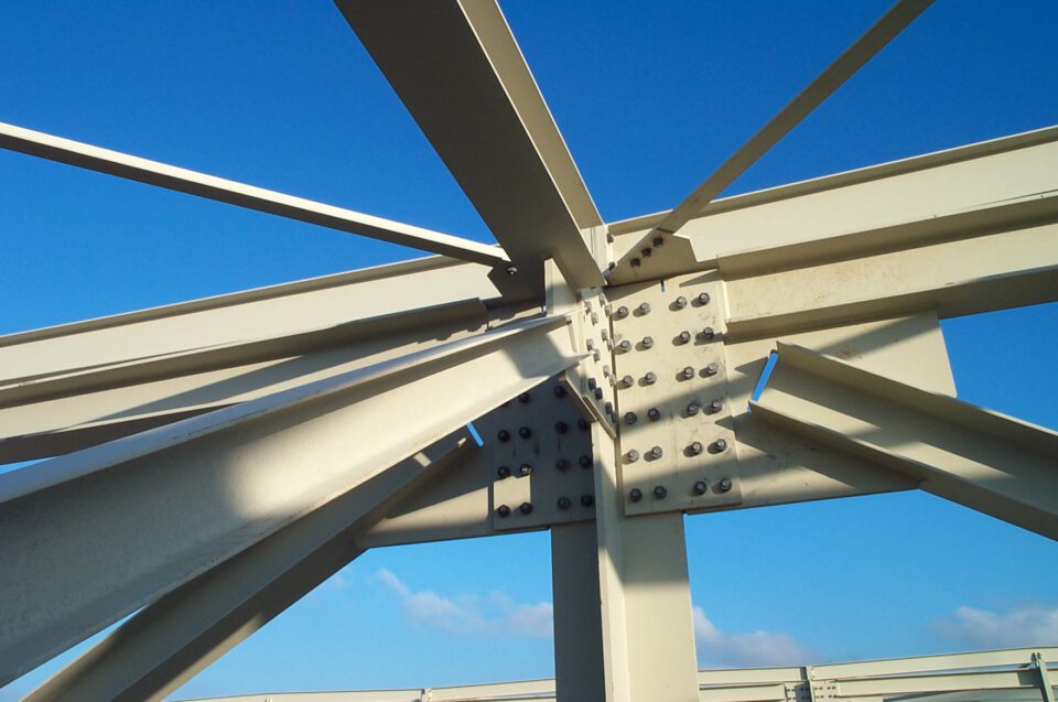 Nahaufnahme einer Stahlkonstruktion mit mehreren Trägern, die an einem zentralen Knotenpunkt verschraubt sind, vor klarem blauen Himmel.