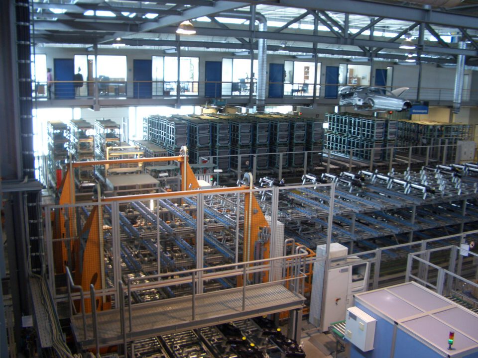 Innenansicht einer großen Industriehalle mit mehreren Ebenen, Metallstrukturen, Lagerregalen mit verschiedenen Materialien und Maschinen; auf der oberen Ebene rechts steht ein Auto, daneben Fenster und Türen zu weiteren Räumen oder Büros.