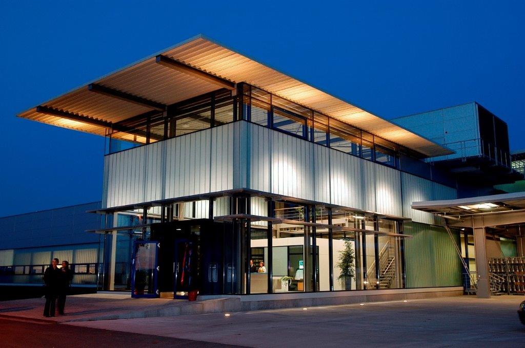 Modernes, zweigeschossiges Gebäude mit großen Glasfenstern und Flachdach, beleuchtet durch Außenlichter, mit zwei Personen auf der linken Seite davor, gelegen in einem gewerblich-industriellen Umfeld.