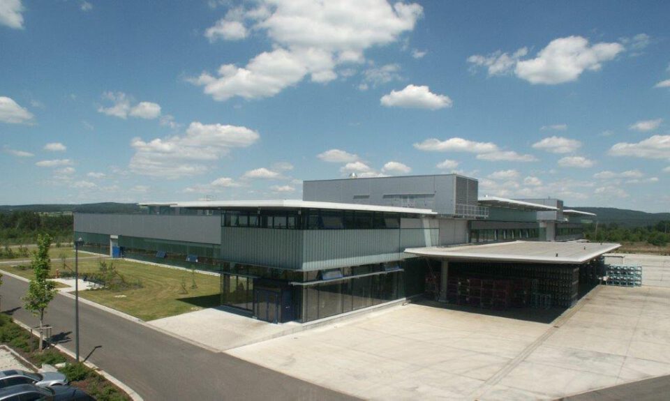Modernes Industrie- oder Geschäftsgebäude mit großem Parkplatz und umliegender Begrünung unter klarem Himmel mit vereinzelten Wolken.