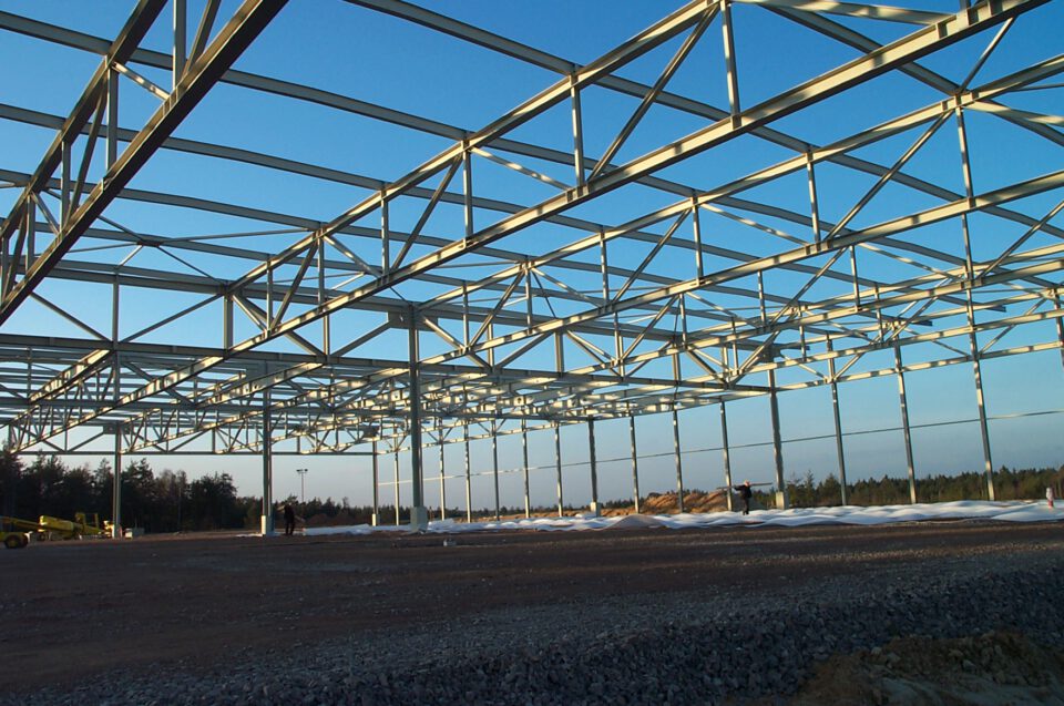 Stahlkonstruktion eines großen Gebäudes im Bau mit zahlreichen vertikalen und horizontalen Trägern in Gitteranordnung sowie diagonalen Verstrebungen, auf einem mit Kies bedeckten Boden und mit Baumaterialien und Geräten im Hintergrund unter klarem Himmel mit wenigen Wolken.
