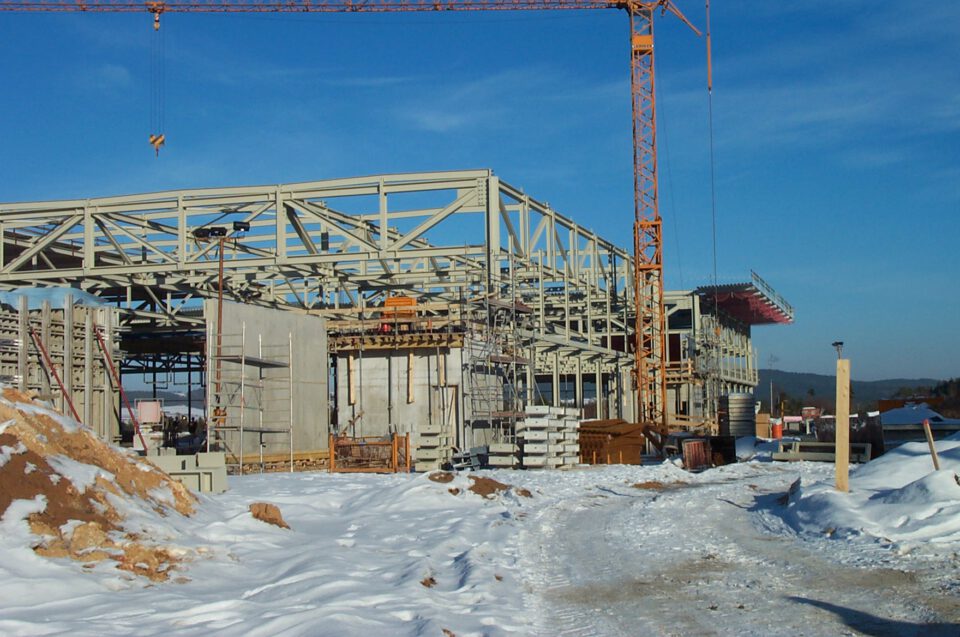 Verschneite Baustelle mit einem teilweise errichteten Gebäude aus Metallträgern, Gerüsten und bereits vorhandenen Betonwänden, umgeben von Baumaterialien und Geräten; ein großer Kran ragt auf der rechten Seite in den Himmel.