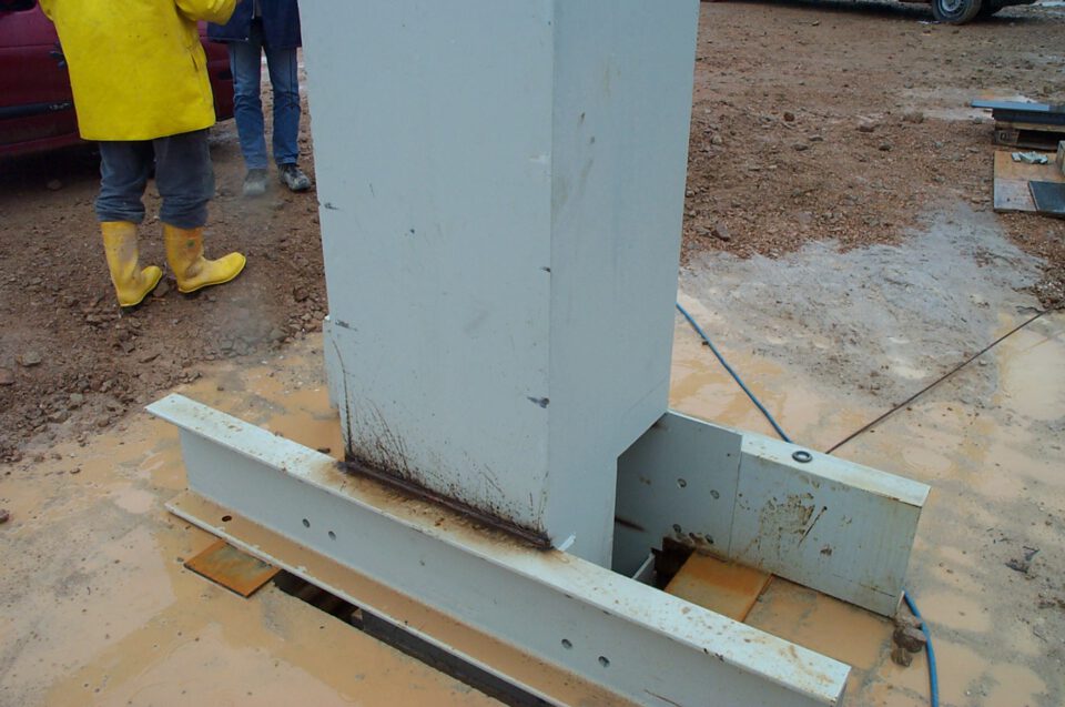 Baustelle mit der Montage einer großen Stahlstütze, die mit Bolzen in einem Fundament verankert ist, umgeben von Metallträgern; im Hintergrund zwei Personen, eine in gelbem Regenmantel und Gummistiefeln, die andere in blauer Jeans, auf schlammigem Boden mit Pfützen.
