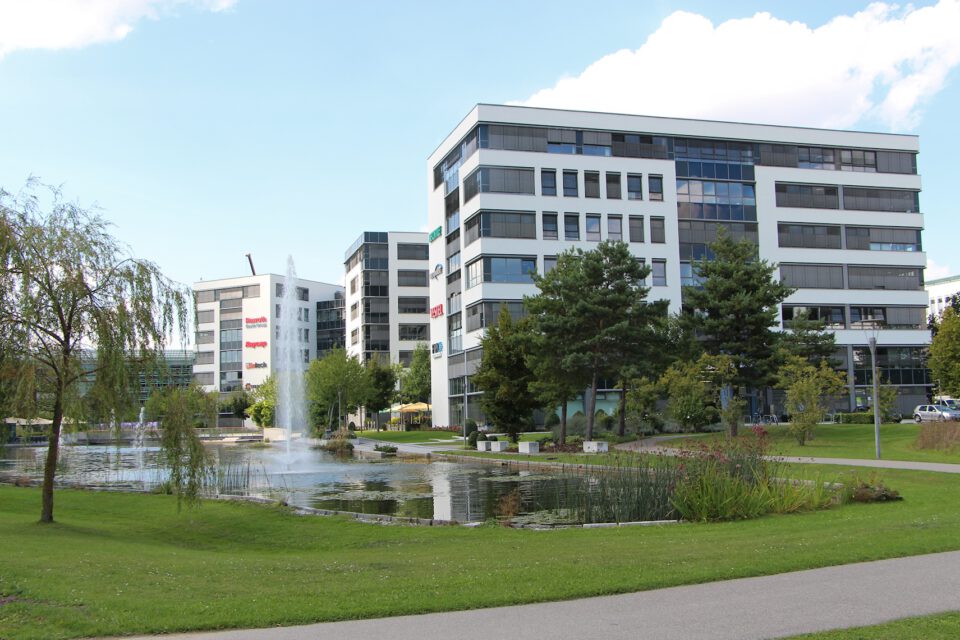 Ein moderner Bürokomplex mit mehreren mehrstöckigen Gebäuden, großen Fenstern und Firmenlogos ist von einer gepflegten Landschaft mit Teich, Springbrunnen, Bäumen und Grünflächen umgeben.