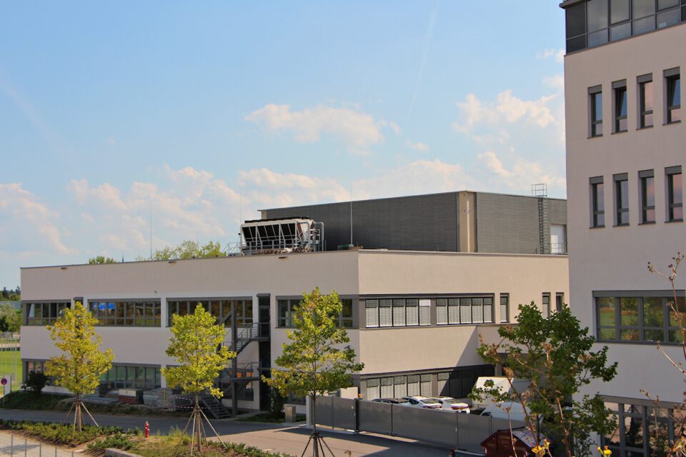 Ein modernes, mehrstöckiges Gebäude mit großen Fenstern und Flachdach steht hinter mehreren Bäumen; rechts ist ein weiteres Gebäude teilweise zu sehen, darüber ein klarer Himmel mit einigen Wolken.