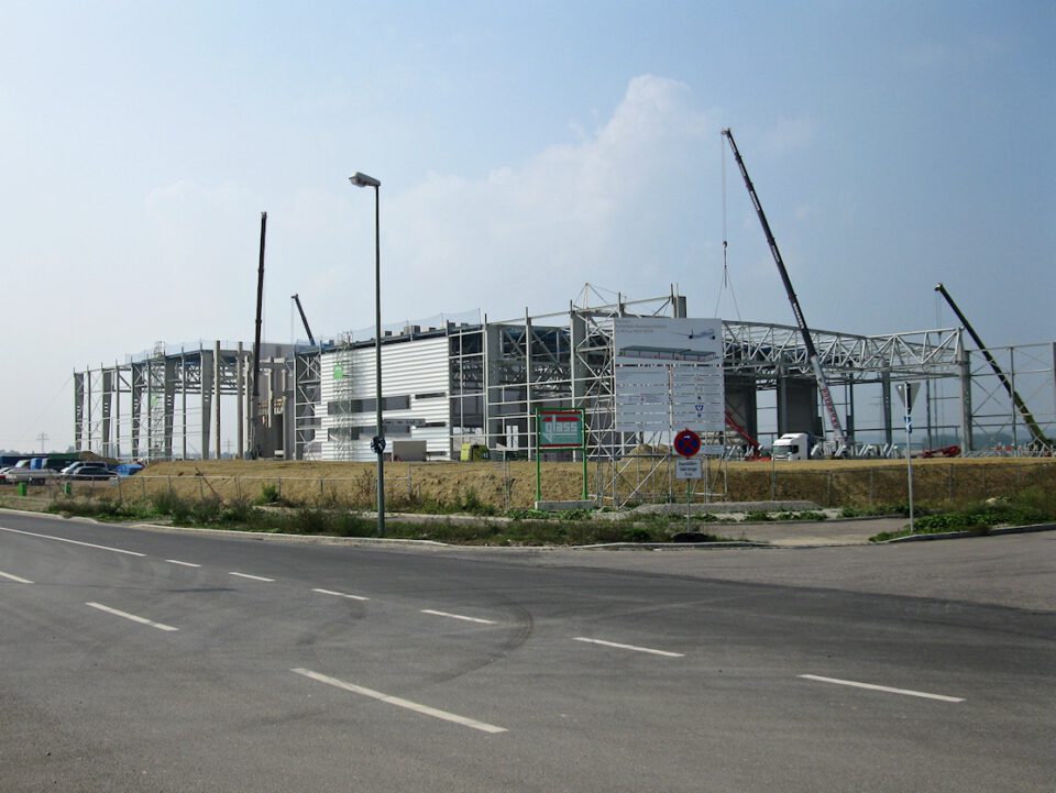 Ein großes Industriegebäude im Bau mit Stahlträgern und mehreren Kränen, die auf laufende Bauarbeiten hinweisen; links sind geparkte Autos zu sehen, im Vordergrund verläuft eine Straße mit einer Straßenlaterne.