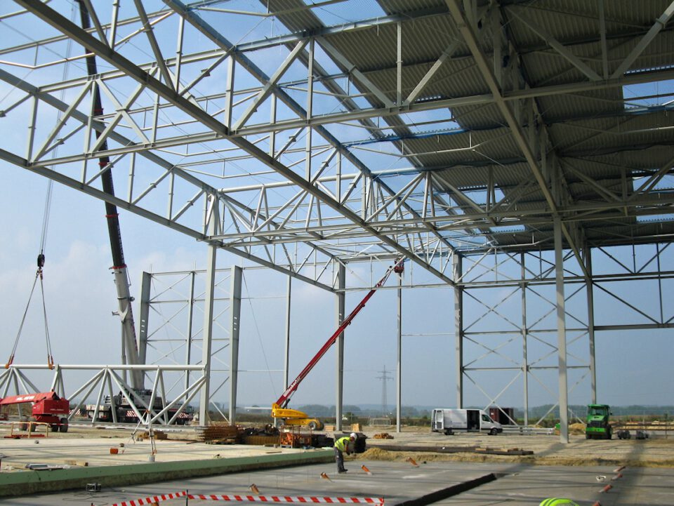 Eine Baustelle mit einem großen Stahlbau, an dem Kräne Stahlträger heben und positionieren; das Gebäudegerüst ist teilweise fertiggestellt, einige Dachabschnitte sind bereits montiert, und rundherum befinden sich Baugeräte, Fahrzeuge und Arbeiter.