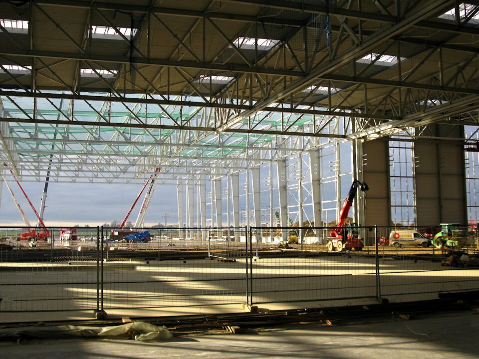 Das Innere eines großen Industriegebäudes im Bau mit Metallträgern, Säulen und mehreren Kränen sowie Baumaschinen im Hintergrund; der Boden ist teilweise fertiggestellt, im Vordergrund steht ein Bauzaun, und Sonnenlicht fällt durch das Dach und wirft Schatten auf den Boden.