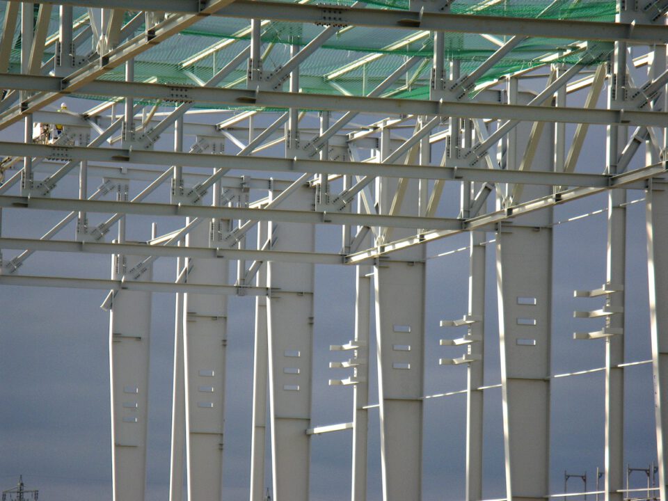 Detailansicht eines Stahlbaus mit vertikalen, horizontalen und diagonalen Trägern, vermutlich Teil eines Gebäudes im Bau; am oberen Rand ist grünes Netzmaterial zur Sicherheit oder zum Auffangen von Bauteilen zu sehen, im Hintergrund ein bewölkter Himmel.