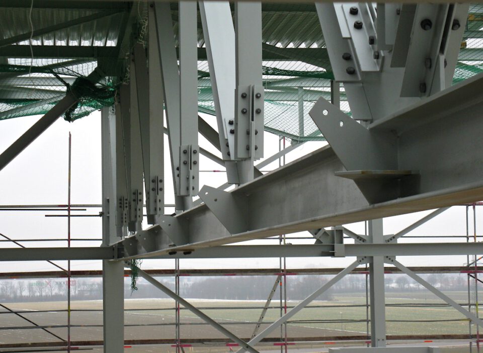 Nahaufnahme eines Stahlbaus im Bau mit mehreren durch Schrauben und Platten verbundenen Trägern und Säulen; im Hintergrund sind grünes Sicherheitsnetz und Gerüste zu sehen, dahinter ein offenes Feld mit einigen Bäumen.