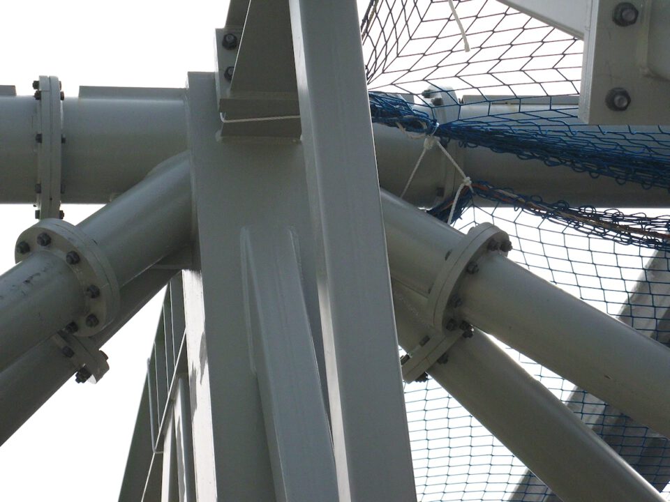 Nahaufnahme eines Tragwerks mit mehreren sich kreuzenden Metallträgern und zylindrischen Rohren, die durch Schrauben verbunden sind; im Hintergrund ist ein blaues Sicherheitsnetz am Bauwerk befestigt.
