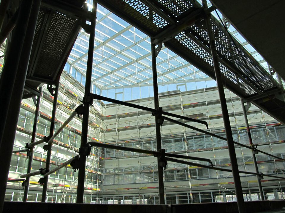 Das Innere eines Gebäudes im Bau oder in Renovierung mit Gerüsten, Metallträgern und Plattformen in einem gitterartigen Aufbau; das mehrstöckige Gebäude verfügt über Glasfenster und ein Glasdach, durch das Tageslicht einfällt.