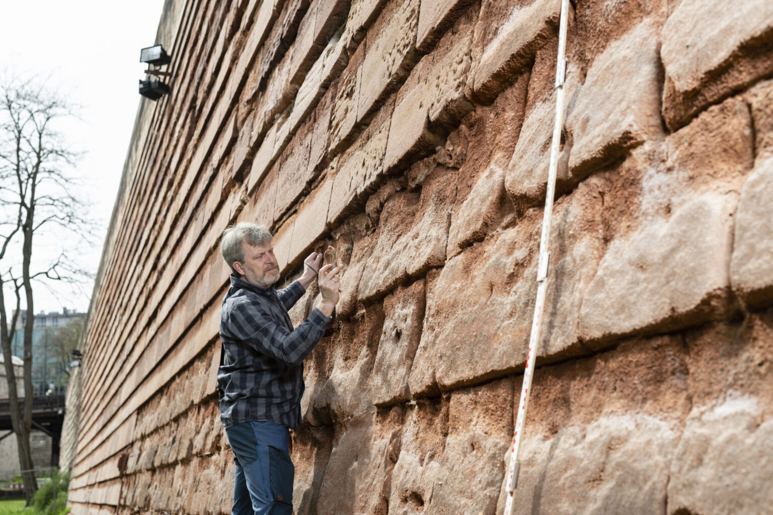 Eine Person in kariertem Hemd und Jeans untersucht die Nürnberger Stadtmauer aus rötlich-braunen Sandsteinblöcken; daneben lehnt ein langer Messstab, im Hintergrund sind Bäume und ein weiteres Bauwerk zu sehen.