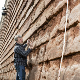 Eine Person in kariertem Hemd und Jeans untersucht die Nürnberger Stadtmauer aus rötlich-braunen Sandsteinblöcken; daneben lehnt ein langer Messstab, im Hintergrund sind Bäume und ein weiteres Bauwerk zu sehen.