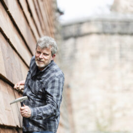 Eine Person mit kariertem Hemd inspiziert die Nürnberger Stadtmauer und untersucht die Steinstruktur mit einem Hammer; im Hintergrund sind weitere Teile der historischen Befestigungsanlage zu sehen.