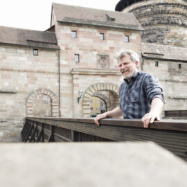 Eine Person steht auf einer Holzbrücke vor der Nürnberger Stadtmauer mit steinernem Mauerwerk, einem Torbogen und Fenstern; im Hintergrund ist ein großer runder Turm zu sehen.